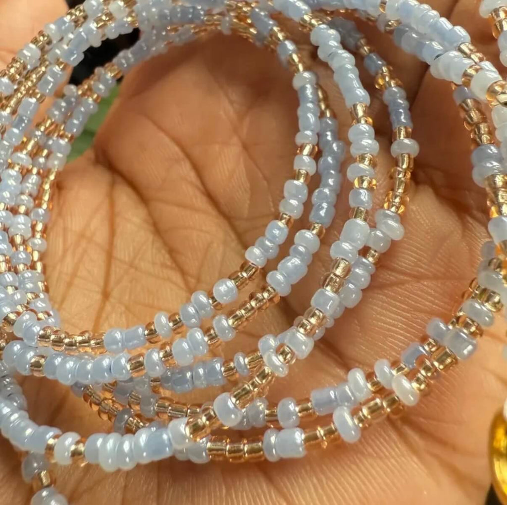 A close-up of multiple beaded waist beads draped over a hand. The bracelets are made of small light blue beads separated by gold beads.